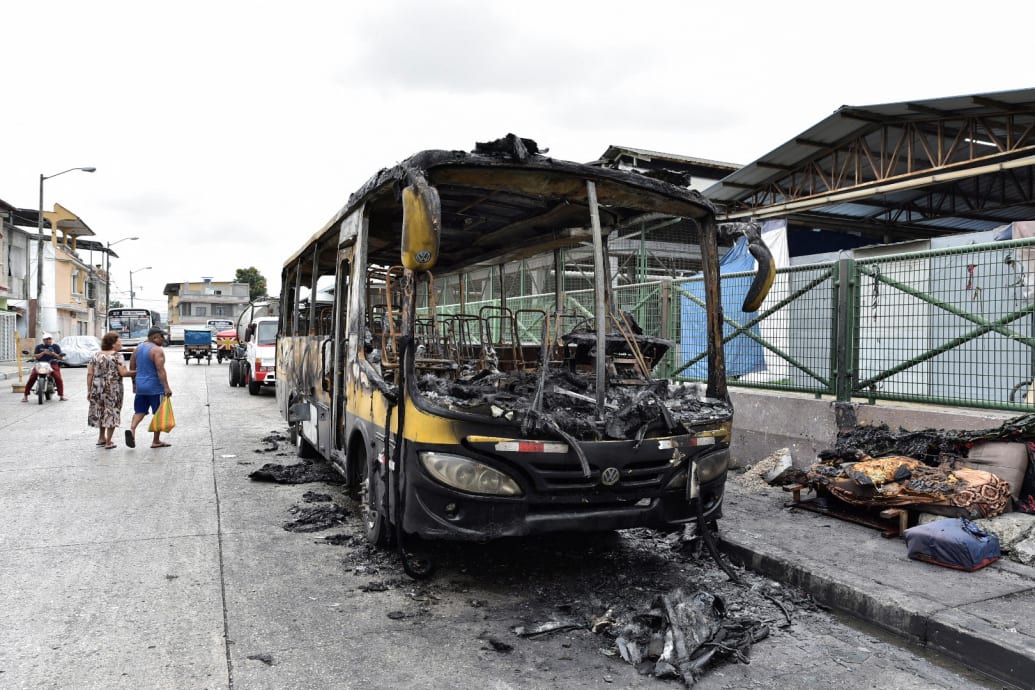 People walk past the remains of a public bus that was set on fire.