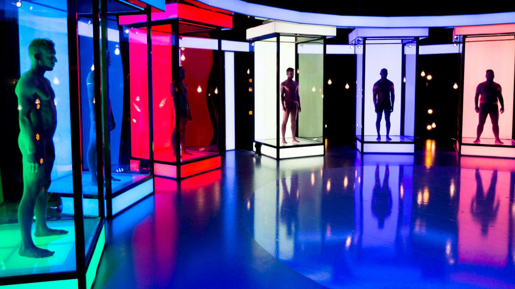 Six naked men stand in dimly lit, neon-colored boxes.