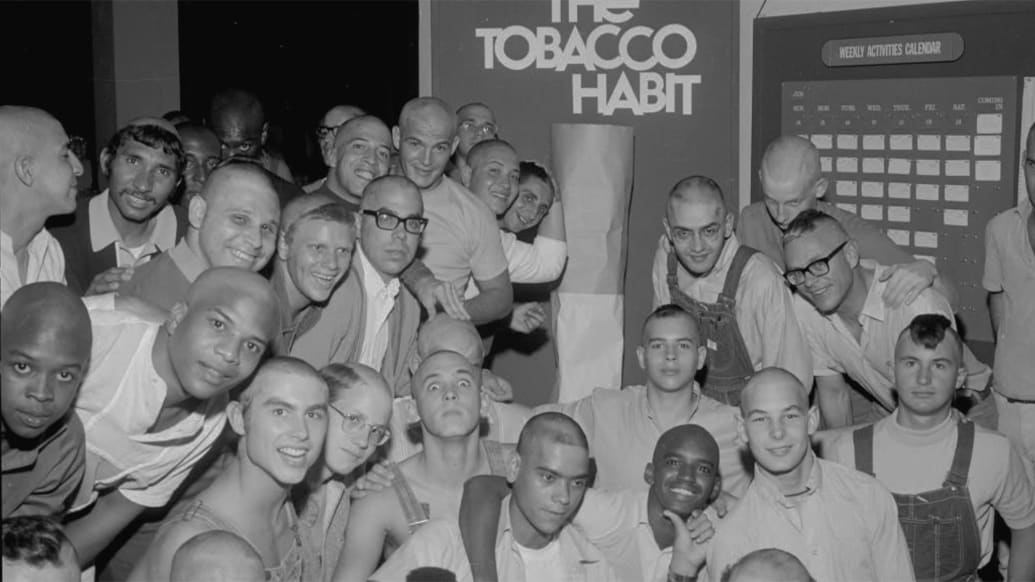 Synanon members standing under sign “Synanon Crushes the Tobacco Habit” in Born in Synanon.