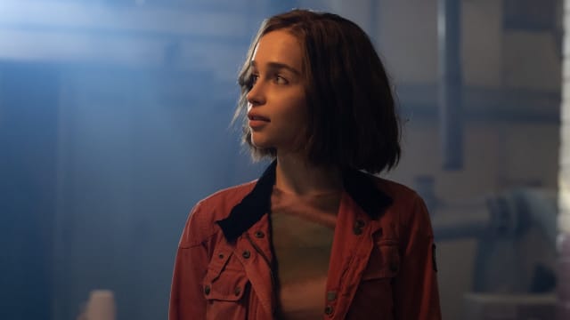 Emilia Clarke looks off into the distance, while wearing a red jacket in a dark room.