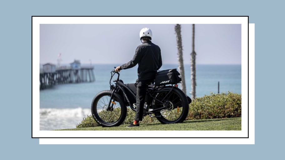 A person wearing a helmet standing beside a black electric bike overlooking the ocean with a pier and palm trees in the background.