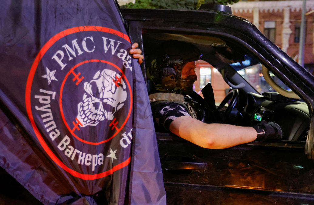 An image of a private mercenary sits in a car next to a Wagner mercenary group flag.