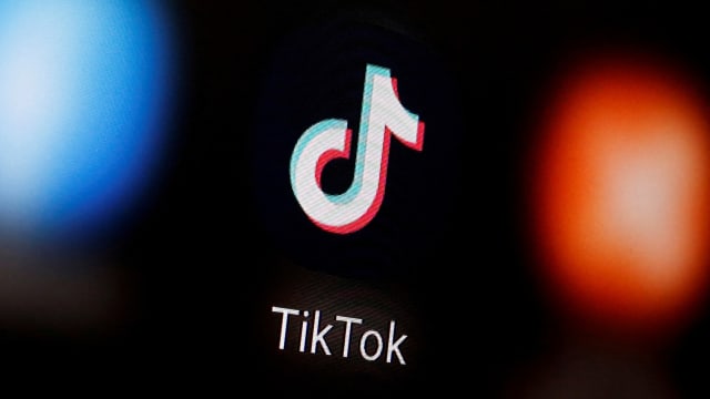 A TikTok logo is illuminated on a black screen with blurred apps on either side of it.