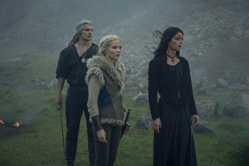 Henry Cavill, Freya Allan, and Anya Chalotra stand in a field from a scene in the third season of The Witcher.
