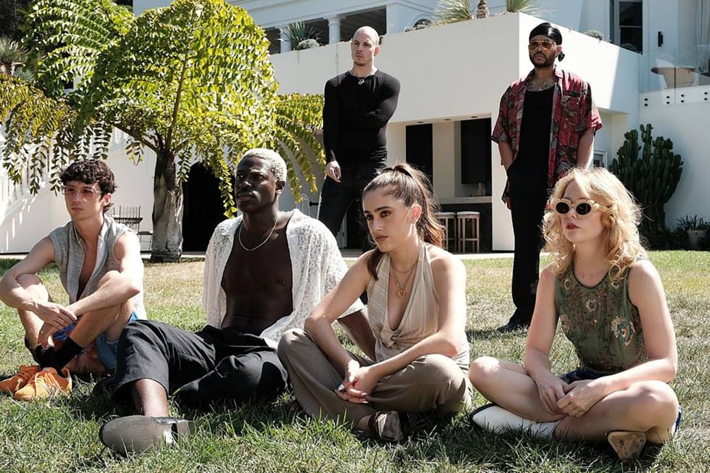 Troye Sivan, Moses Sumney, Mitch Modes, Rachel Sennott, Abel "The Weeknd" Tesfaye, and Suzanna Son sit on the grass in the backyard of a mansion in a scene from The Idol.