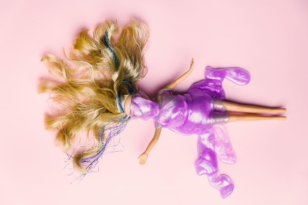 A doll laying on a pink background and covered with purple shiny gel.