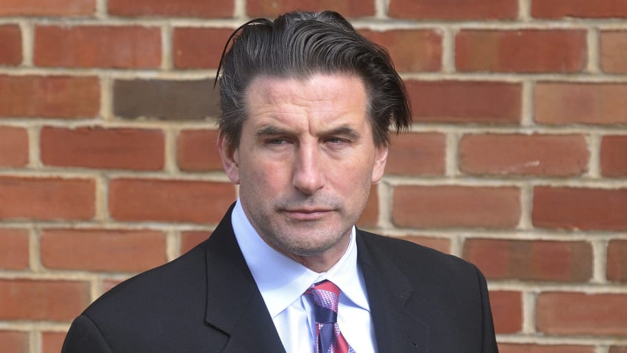 Billy Baldwin wears a suit in front of a brick wall