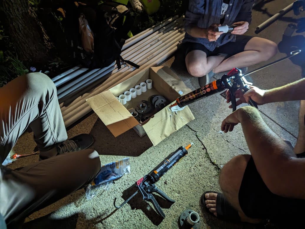 Three members of the People's CDOT prepare for the installation on a sidewalk. One is holding a pipe fitting in their hand, while another holds a caulk gun.