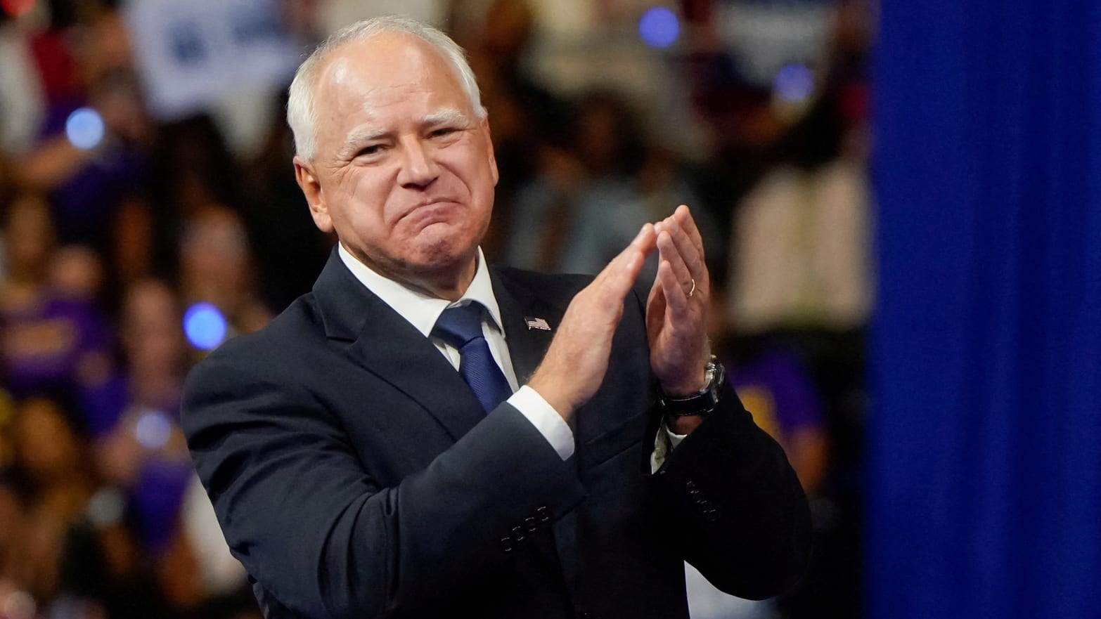 Tim Walz smirks and claps on stage at a rally.