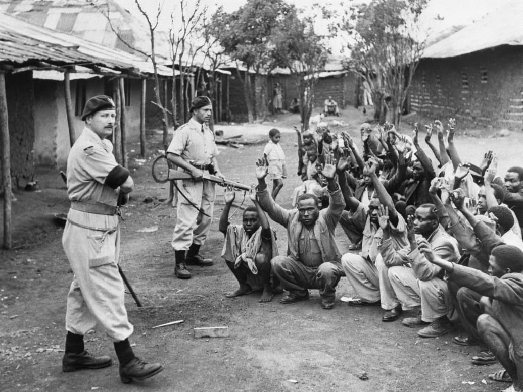 British policemen hold men from the village of Kariobangi at gunpoint while their huts are searched for evidence that they participated in the Mau Mau Rebellion.