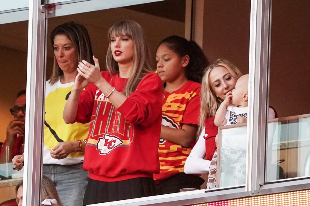 A photograph of Taylor Swift wearing a vintage Kansas Chiefs Jacket during a Chiefs game.