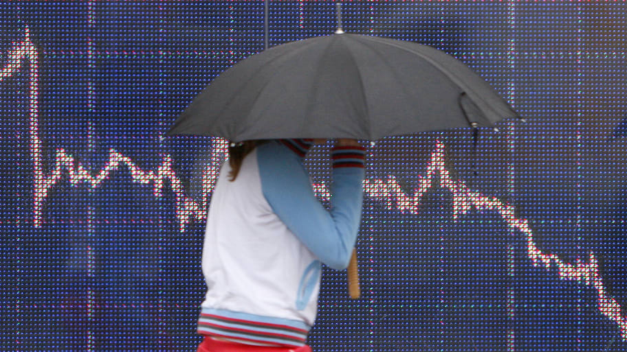 A woman shelters from the rain as she passes a screen showing trading market graphs