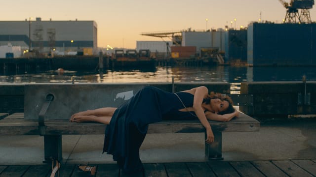 A woman lies on a bench in a blue dress near the water.