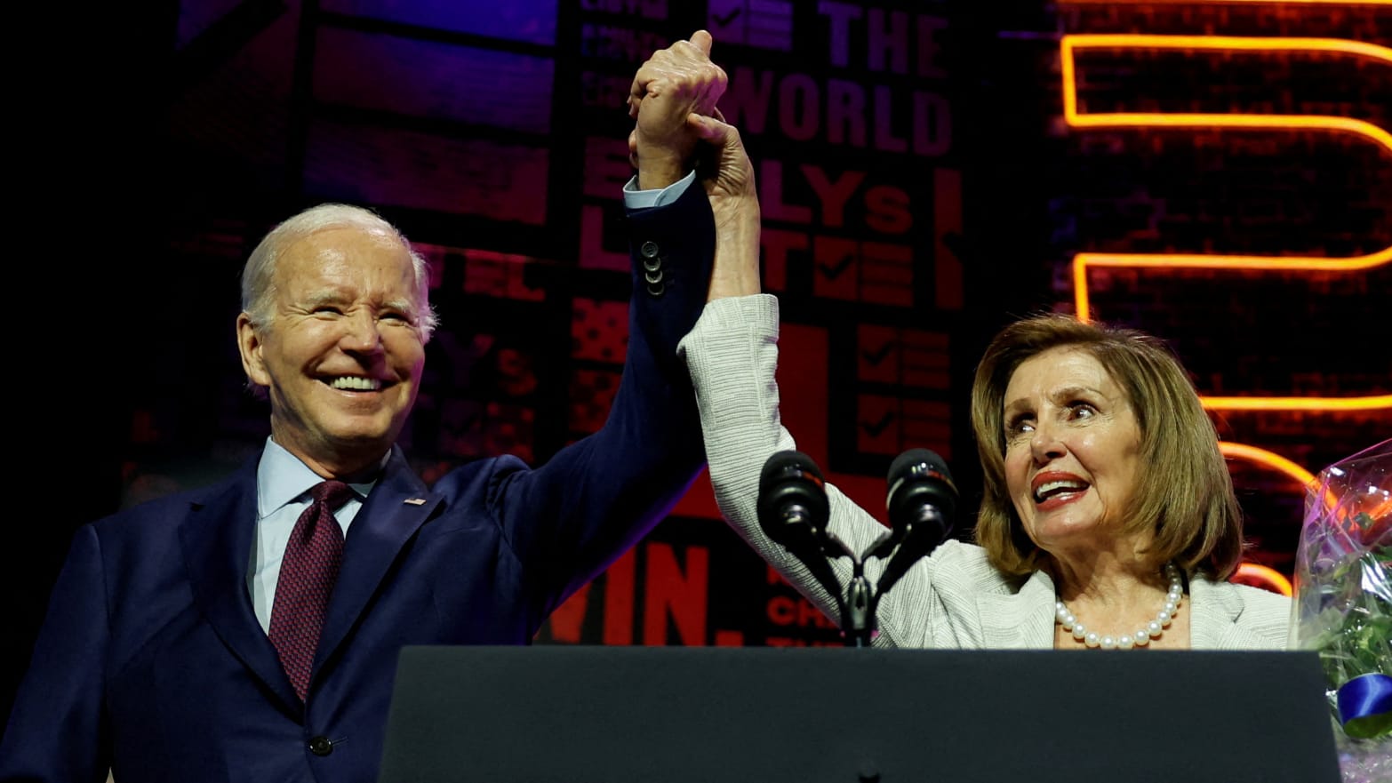 Joe Biden and Nancy Pelosi raise their arms while holding hands on stage 