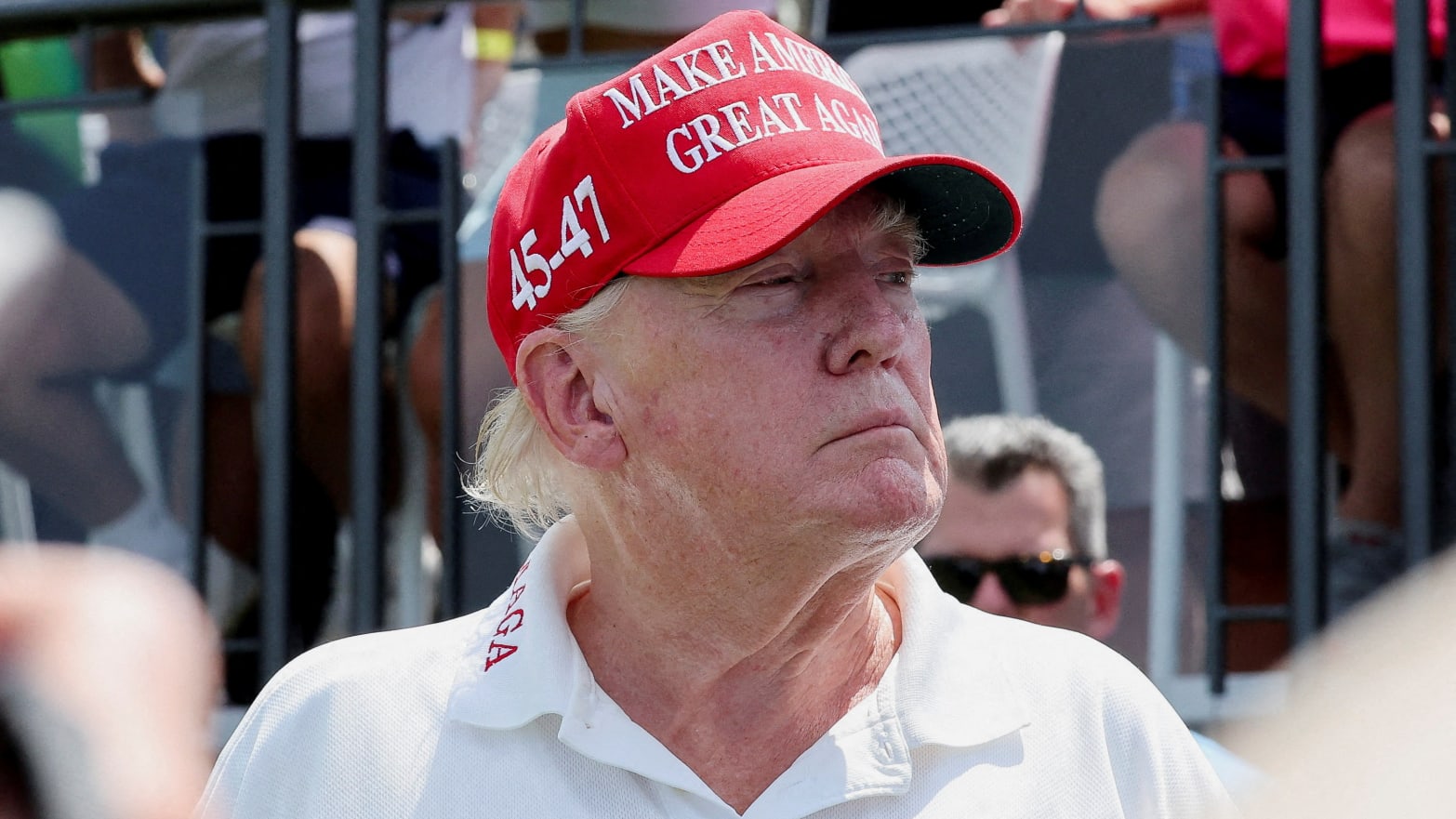 Donald Trump at a golf course wearing a red MAGA hat and a white polo shirt.