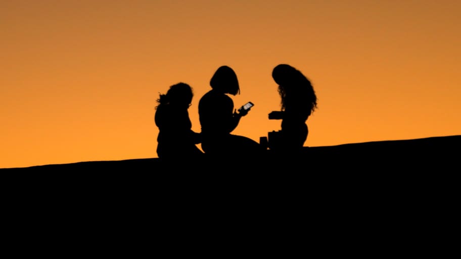 Teen girls look at their phones on a hillside during sunset.