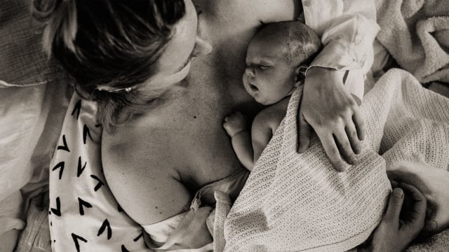 A photograph of a mother with a baby laying on her chest in bed at home.