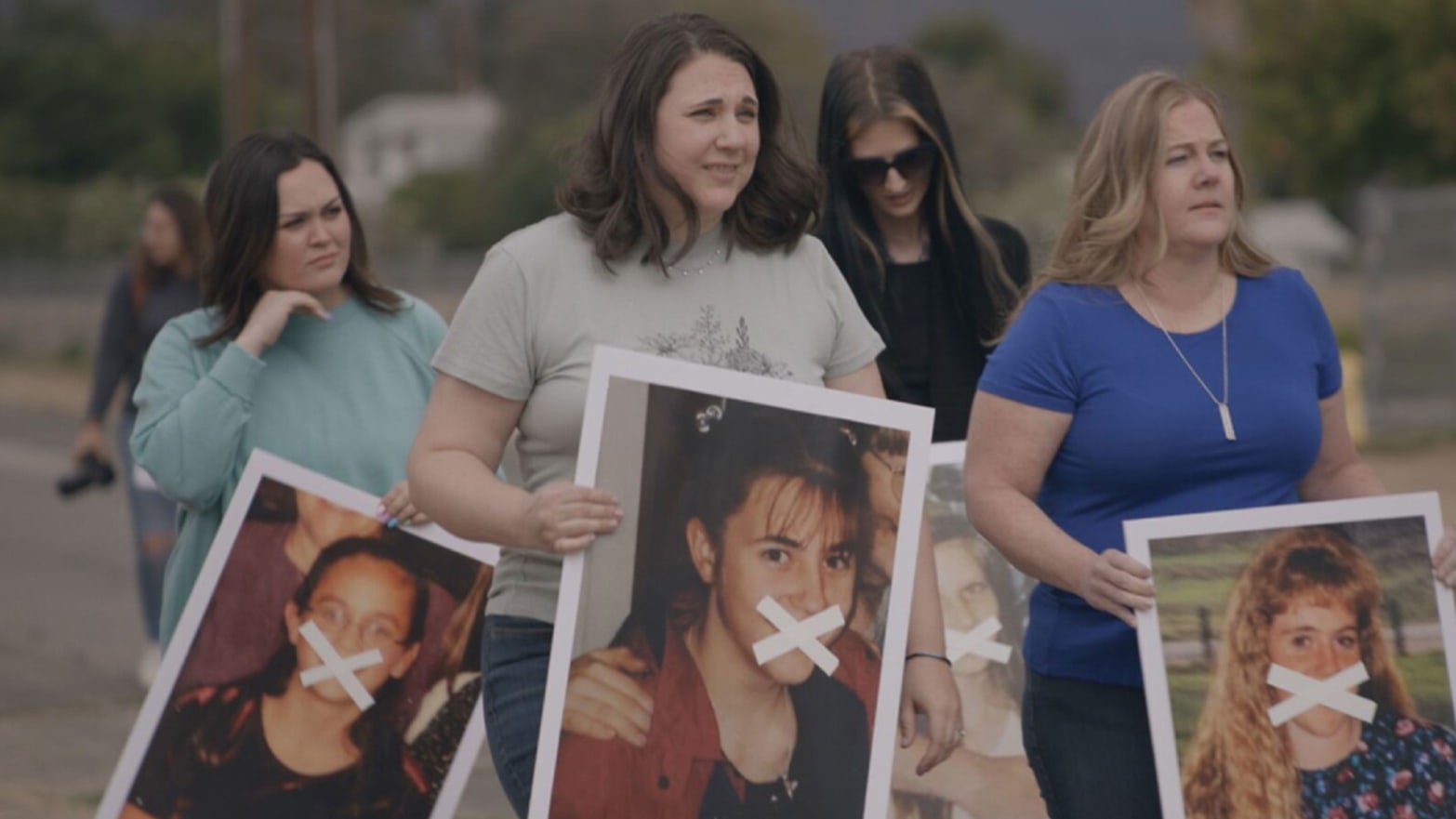 Women march holding pictures of their younger selves with x's crossed over their mouths in a still from 'Let Us Prey'