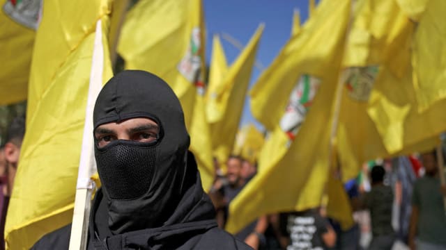 A photo of a man in a black mask in front of a crowd of Palestinians waving the yellow Fatah flag at a protest