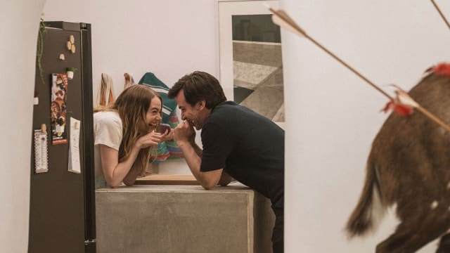 Emma Stone and Nathan Fielder hold hands and laugh over a counter in a still from 'The Curse'