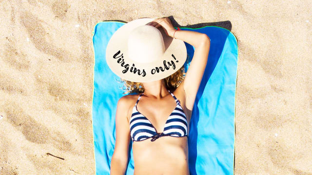 A woman on the beach lays on a towel with a hat that says ‘Virgins Only’