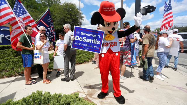 A person wearing a mouse costume holds a Governor Ron DeSantis poster
