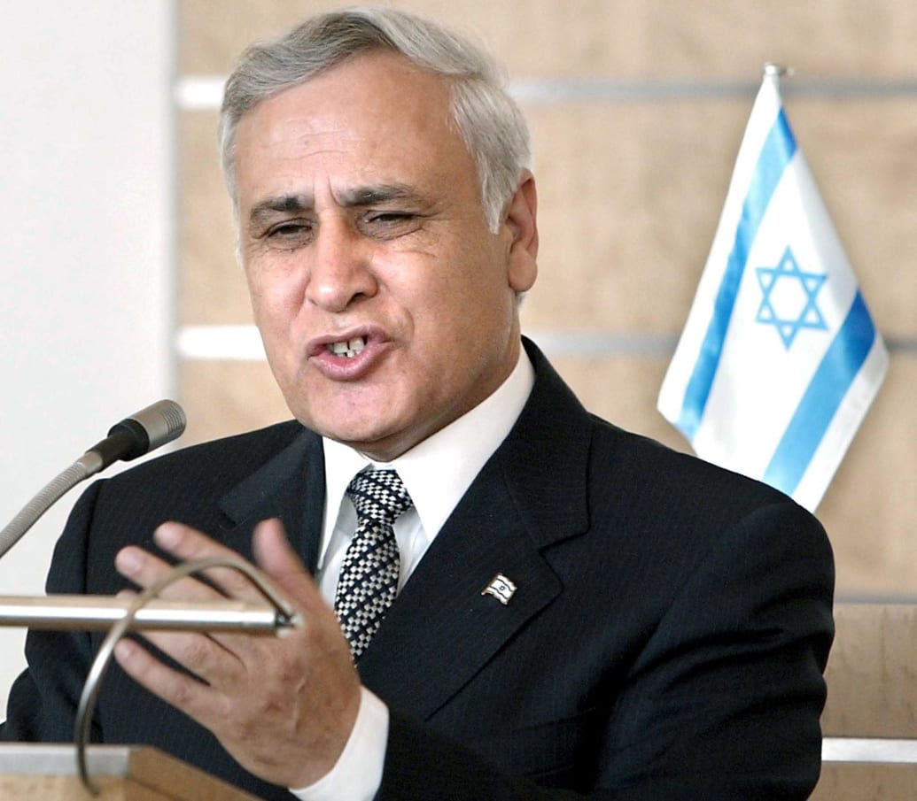 Moshe Katsav speaks at a podium with an Israeli flag in the background.