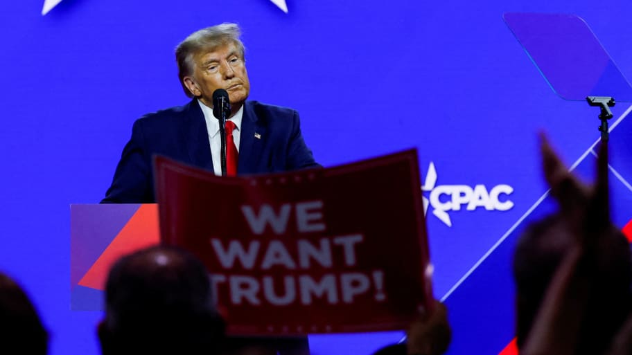 Donald Trump speaks at a conference while an audience members holds a sign that reads, “WE WANT TRUMP!”