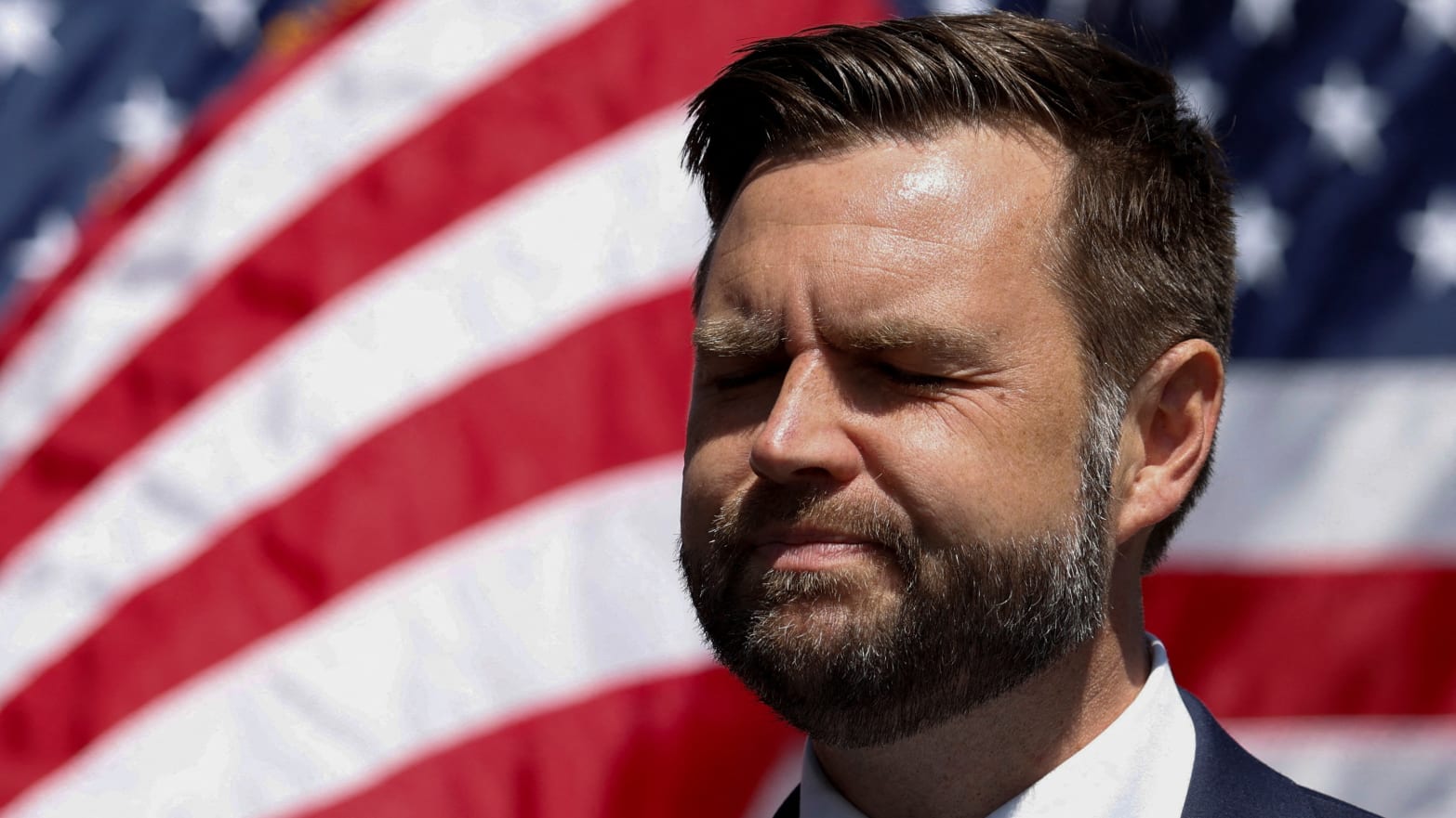 JD Vance closes his eyes on stage with American flags in background.