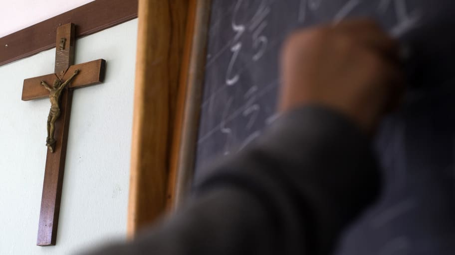 A crucifix is seen on a wall as a student writes on a blackboard