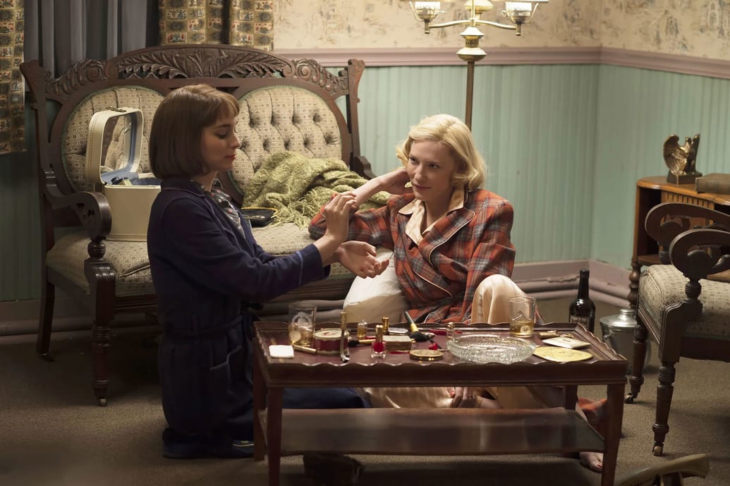 Rooney Mara and Cate Blanchett sit in a living room trying on perfume in a still from 'Carol'