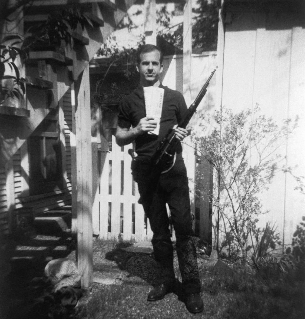 Lee Harvey Oswald holds a Mannlicher-Carcano rifle and newspapers in a backyard.
