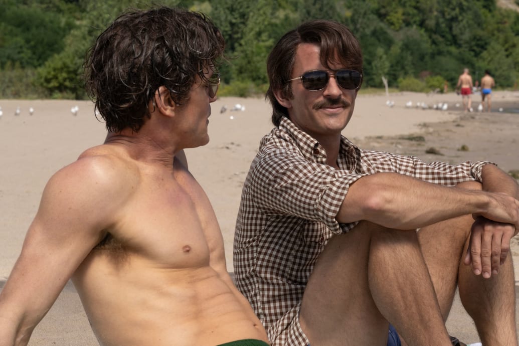 Matt Bomer and Jonathan Bailey sit shirtless on a beach in a still from ‘Fellow Travelers’