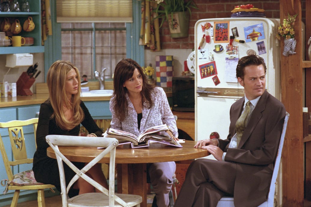 Jennifer Aniston, Courteney Cox and Matthew Perry sit together at a table on the set of "Friends".