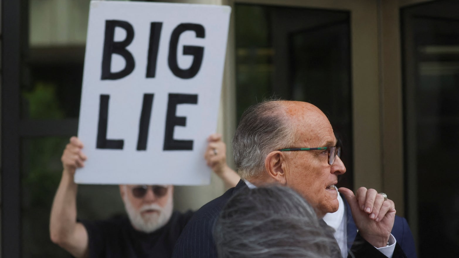 Rudy Giuliani, wearing a suit, leaves a courthouse flanked by protesters, including one holding a sign that says, “BIG LIE.”