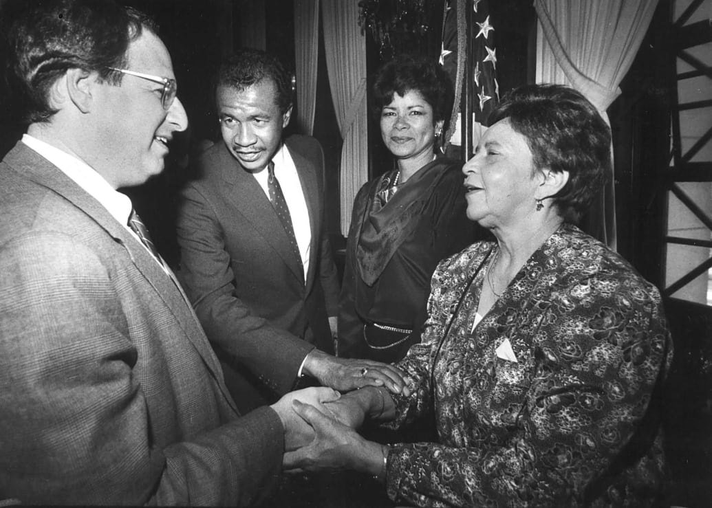 A black and white photo shows Mark Wolf shaking hands with Jacquline Budd while Wayne Budd and Octavia Budd look on.