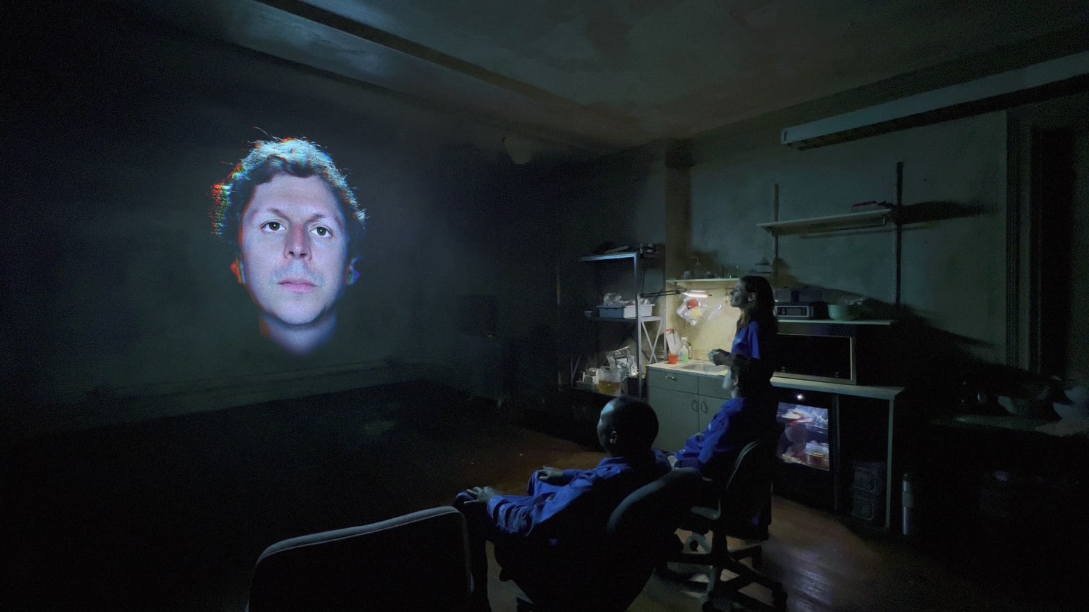 Michael Cera’s disembodied head on a screen, staring straight ahead, as three people sit in armchairs in a dark room below.