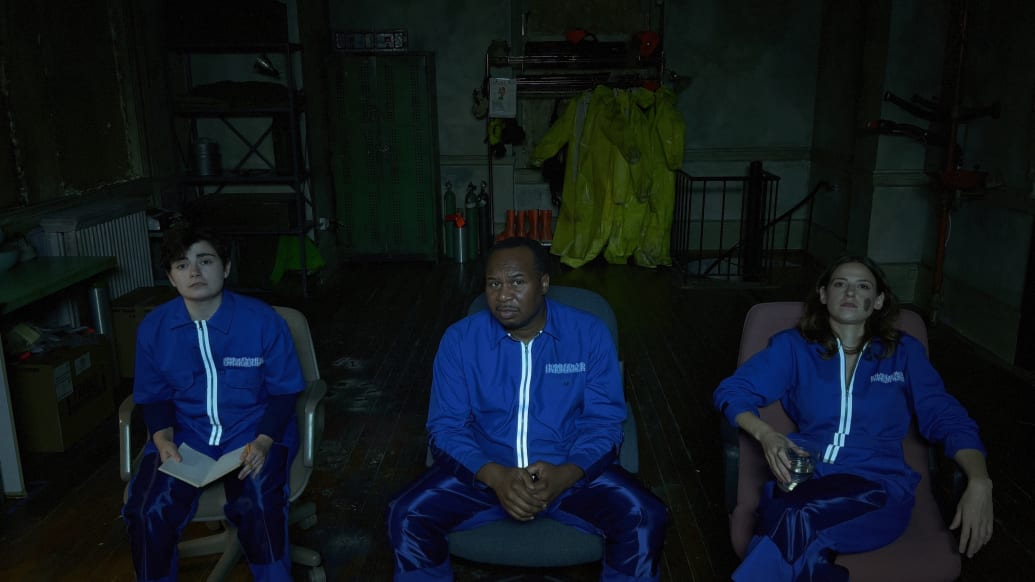 Three people in jumpsuits stare up at a screen, sitting in armchairs.