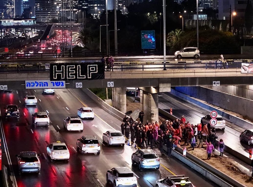 Protestors gather on a highway. A help flag can be seen thrown off an overpass.