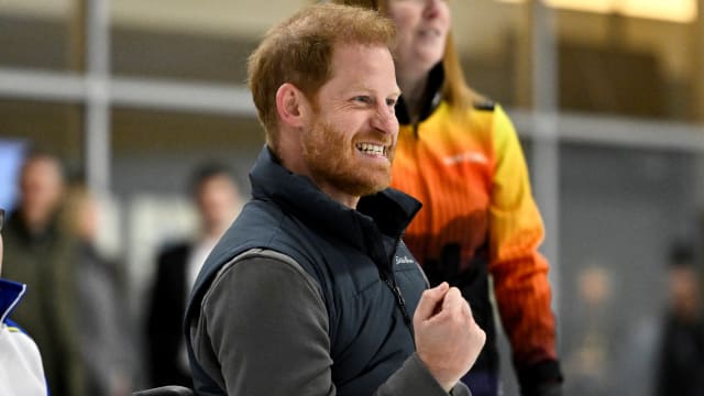 Prince Harry stands with his arm slightly raised while watching a sporting event.
