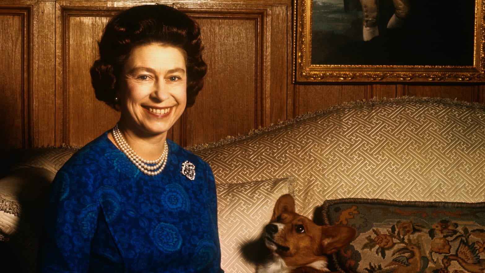 Queen Elizabeth II smiles radiantly during a picture-taking session in the salon at Sandringham House. Her pet dog looks up at her.