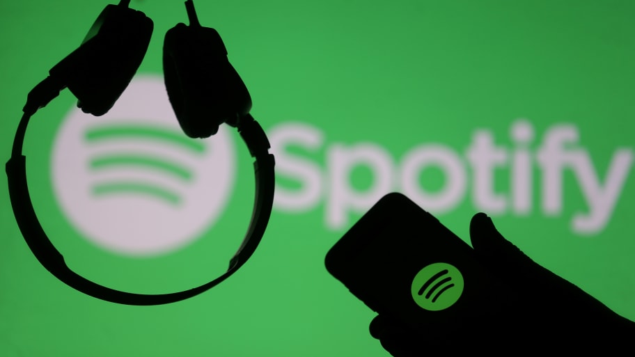 A smartphone and a headset are seen in front of a screen projection of Spotify logo.