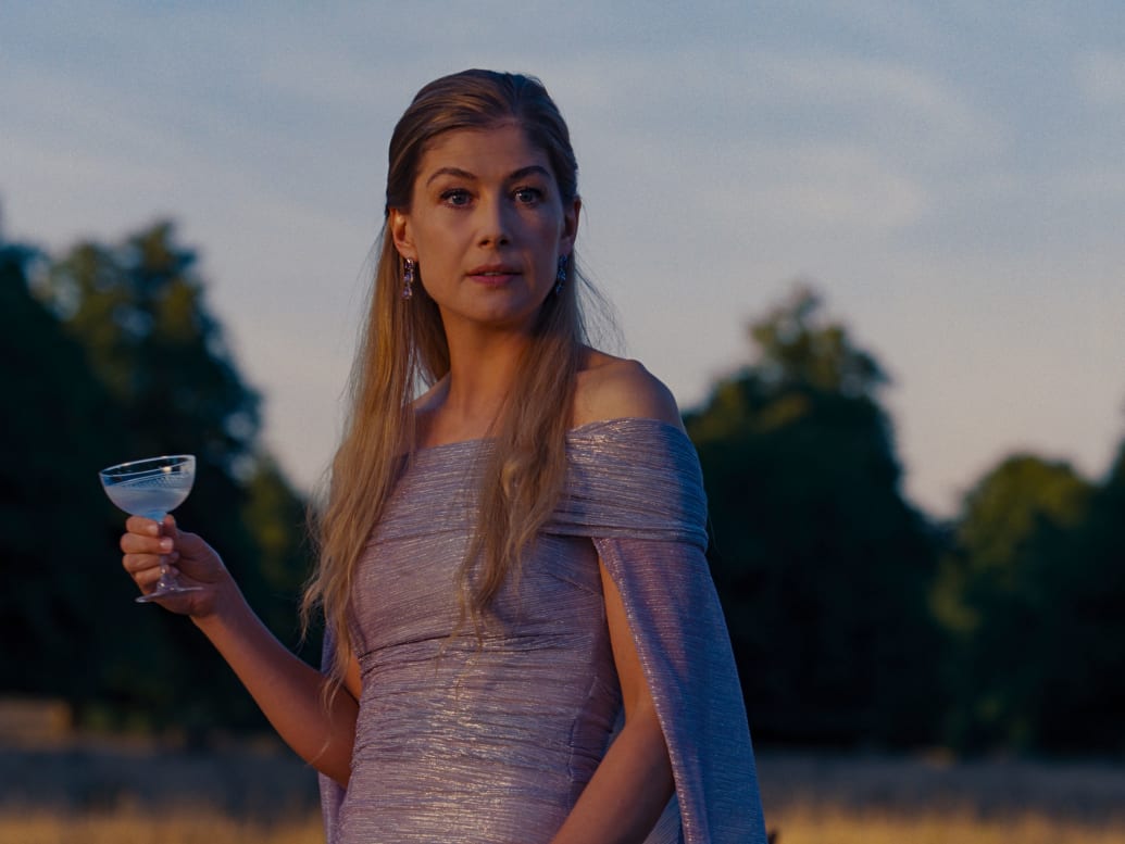 Rosamund Pike in a purple dress holds a cocktail in a still from ‘Saltburn’