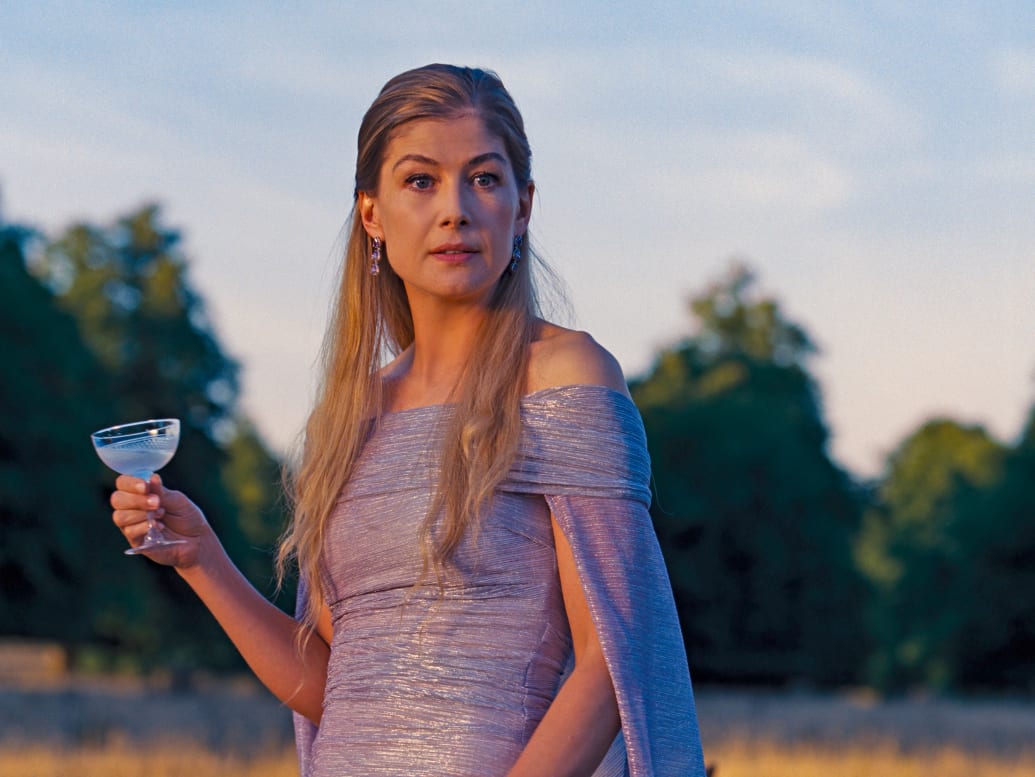 Rosamund Pike wears a purple dress and carries a cocktail outside in a still from ‘Saltburn’