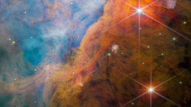  A nebula made of many layers of cloudy, colourful material. The top-left side of the image is brightly lit, filled with wispy, thin material in pale shades of pink and blue. A thick bar of denser, cloudier material crosses diagonally at the bottom right.