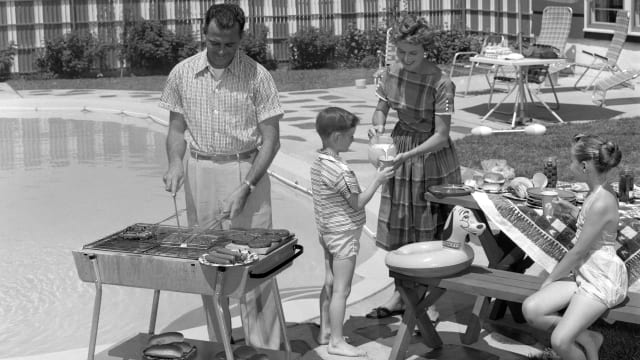 A 1950s family scene of a father grilling by a pool with his wife, son and daughter standing close by. In the background is a ranch-style home.