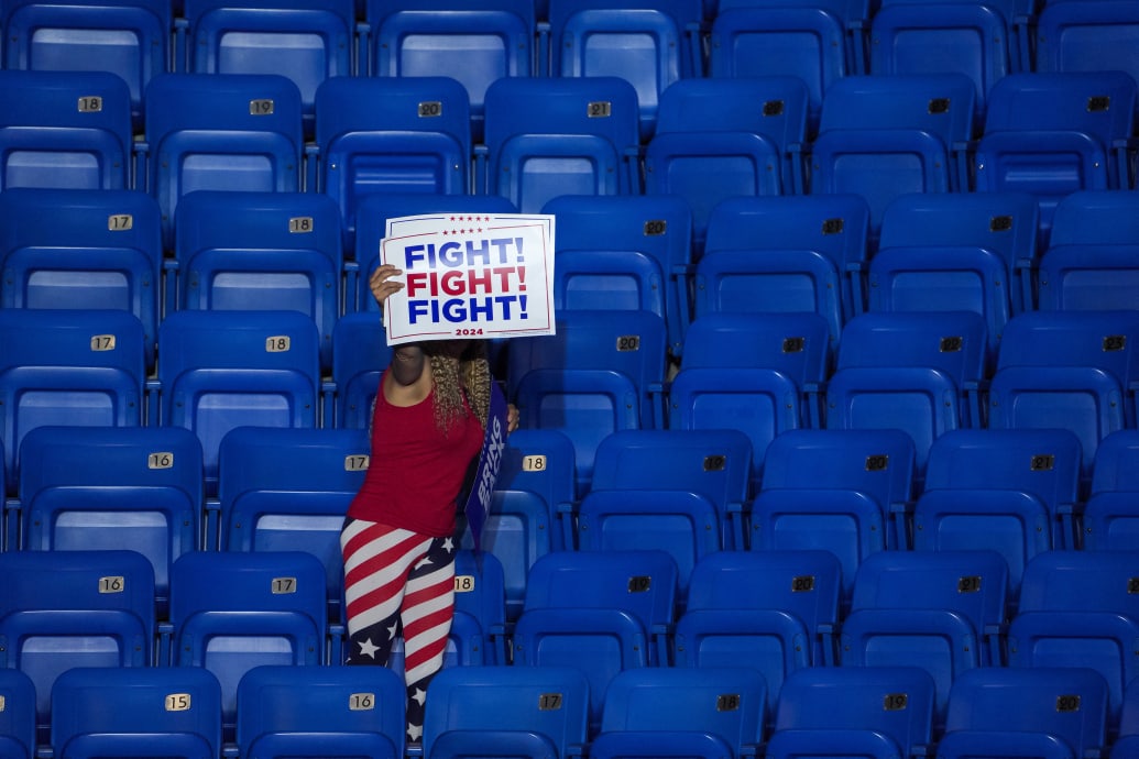 A woman surrounded by empty seats holding up a Trump sign saying “fight.”