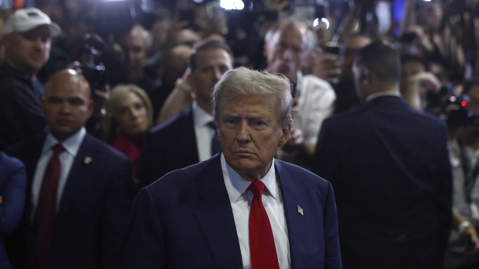 Donald Trump stands withs his back to a busy crowd of people in suits and cameramen, frowning as he looks off into the distance.