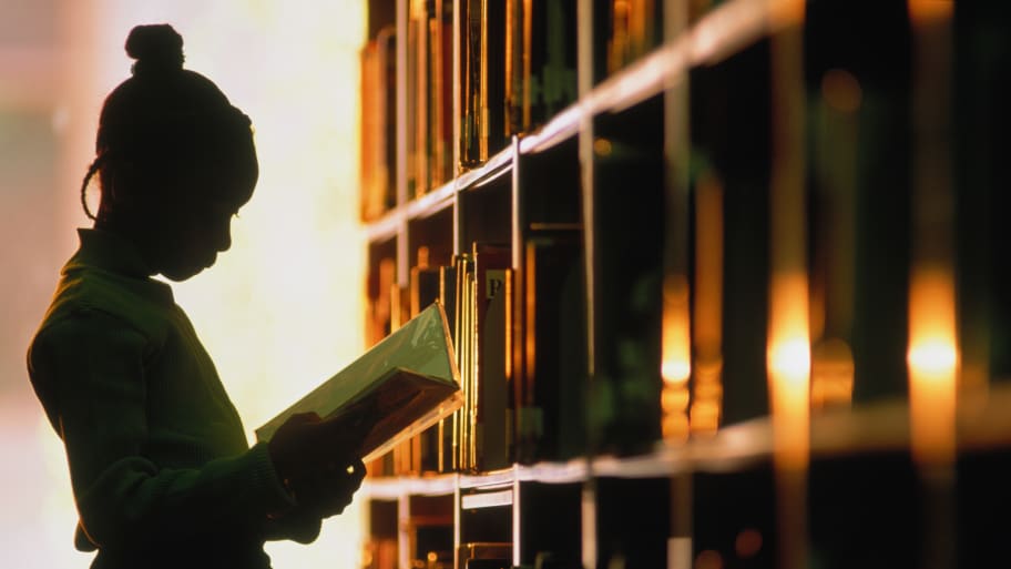 Young school girl stands holding a library book silhouetted next to a library book shelf. 