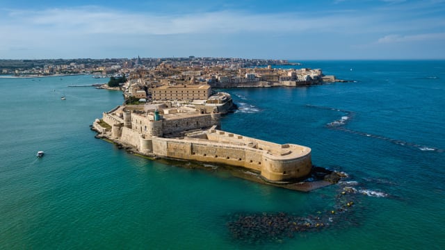 Drone view towards the "crocodile head shaped" Ortygia Island connected to Syracuse City with Maniace Castello at the Eastern Coast of Sicily Island towards the Mediterranean Sea under blue summer sky. 